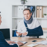 middle aged worker in neck brace with broken arm sitting at table and talking to businessman in blue jacket in office, compensation concept