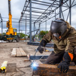 A young  man welder in brown uniform, welding mask and welders leathers, weld  metal  with a arc welding machine at the construction site, blue sparks fly to the sides