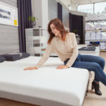 Attractive female customer sitting on a new bed at furniture store, choosing orthopedic mattress to buy