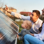 Man shows his family the solar panels on the plot near the house during a warm day